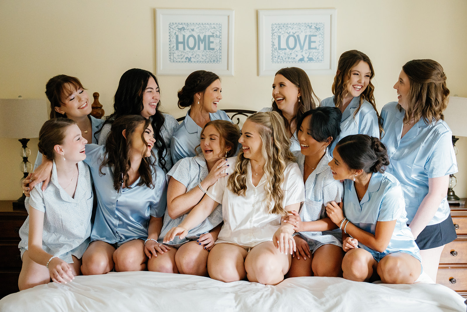 Bride laughing with her bridesmaids in matching blue getting-ready pajamas during wedding morning preparations in a bright, cozy bedroom.